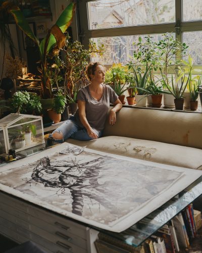 Artist Heidi Jung sits in her home surrounded by plants and her artwork.
