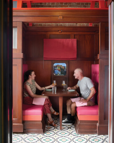 Two guests enjoy drinks in a wood-paneled booth with bright pink seating, patterned tile flooring, and train-inspired details at Rōmyō in Denver's Larimer Square.