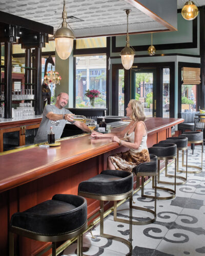 A bartender pours wine for a guest at Rōmyō in Denver, featuring a polished wood bar, black velvet stools, brass pendant lighting, and decorative patterned tile flooring.