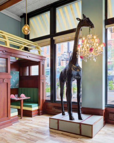 A towering black giraffe sculpture holding a crystal chandelier adorned with pink flowers stands near the windows at Rōmyō in Denver's Larimer Square, showcasing the restaurant's playful interior design.