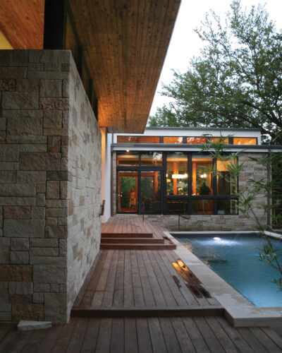 Modern custom home featuring natural stone walls, wood decking, floor-to-ceiling windows, and a swimming pool illuminated at dusk.