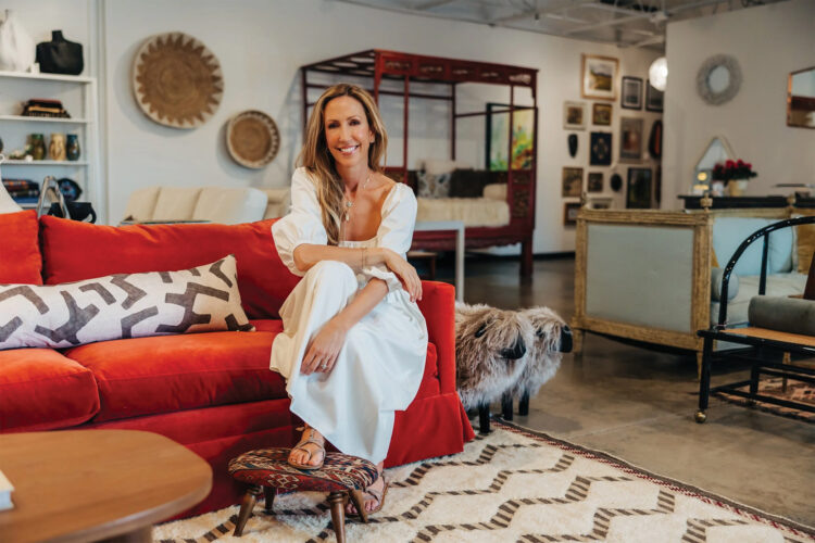 Paula Minnis, founder of The Refinery in Boulder, Colorado, sits on a bright red sofa surrounded by consignment furniture, vintage décor, artwork, and globally inspired home furnishings.
