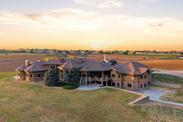 Aerial view of a sprawling luxury home with stone accents, a tiled roof, and expansive grounds surrounded by farmland at sunset.