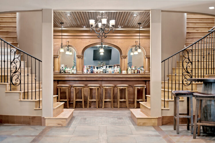 Elegant home bar with wood cabinetry, decorative metal ceiling tiles, chandeliers, and twin staircases featuring wrought-iron railings.